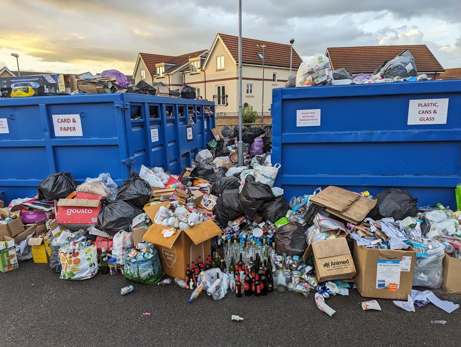 Photo of recycling material piled up around two blue skips.